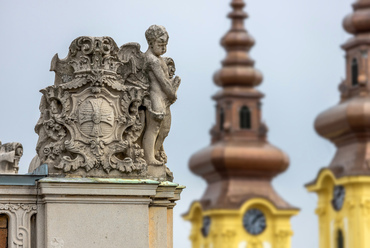 Angyallal díszített ciszterci címer a főgimnázium attikáján. A háttérben a Szent Imre-templom tornyai láthatók